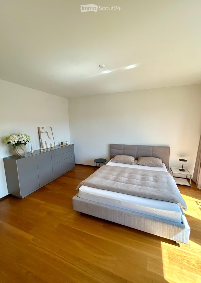 Modern bedroom with wooden flooring, a grey upholstered bed, nightstand, mirror, and decorative vase on a dresser.