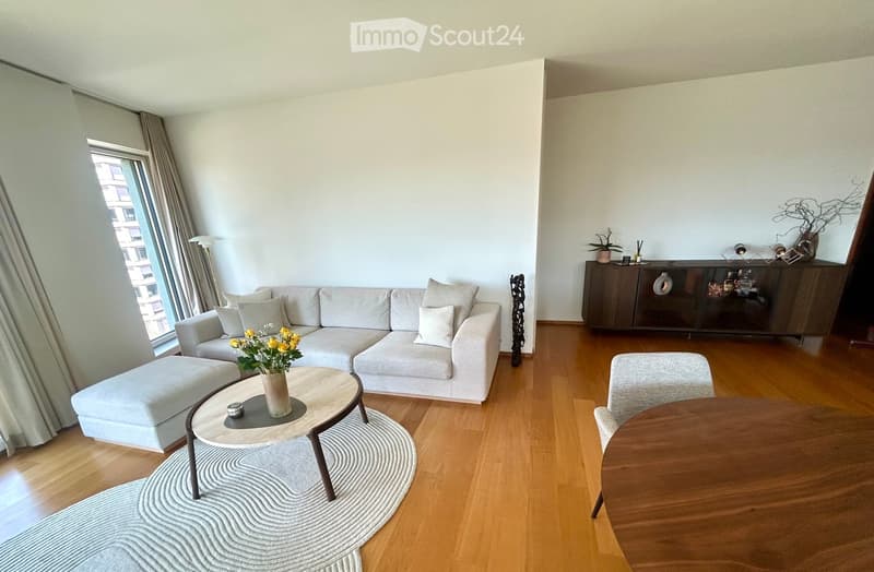 Modern living room with light-colored furniture, including a sectional sofa, coffee table, and dining table, set against wooden flooring and white walls.