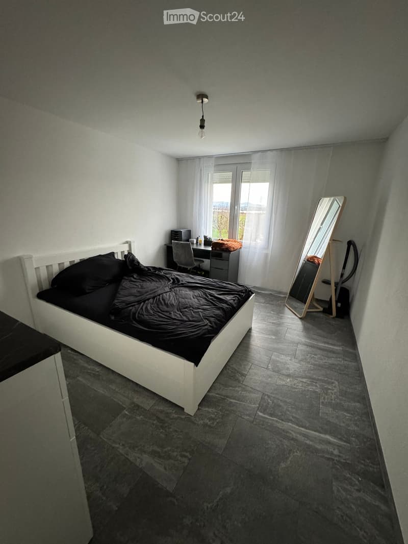 Modern bedroom with a white bed, dark bedding, a large mirror, a desk with a chair, and a lamp hanging from the ceiling.