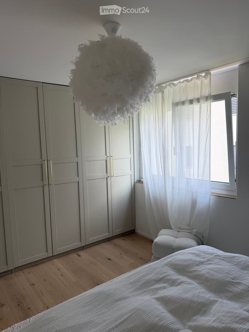 A modern bedroom with a large bed, a wardrobe, a chair, and a decorative light fixture. The room features wooden flooring and large windows with white curtains.