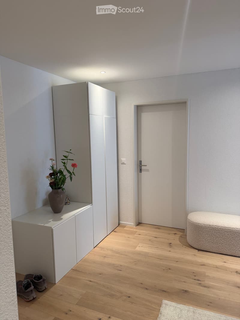 A modern, minimalist hallway with light wooden flooring, white walls, a built-in shoe rack, a small round stool, and a vase with flowers on the shelf.
