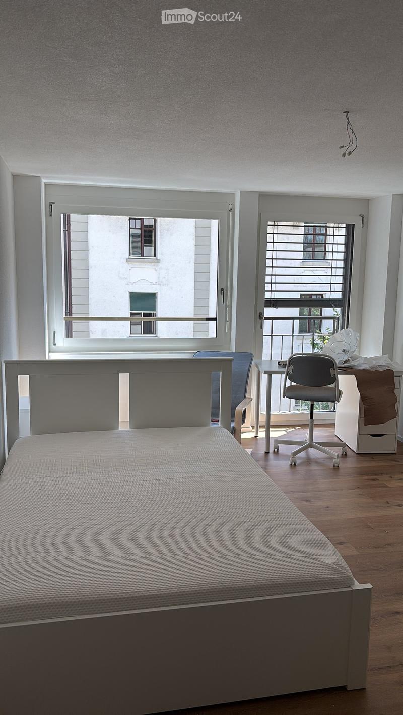 Modern bedroom with a single bed, a small desk with a chair, and a cabinet. Large windows offer a view of the balcony and the street outside.
