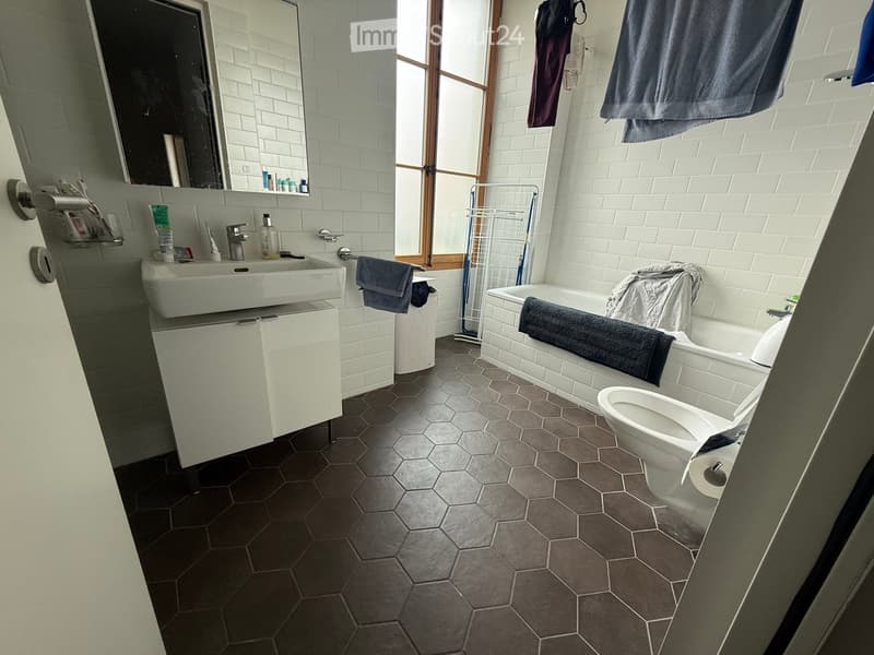 Modern bathroom with white tiles, hexagonal dark grey floor tiles, a white sink with a cabinet, a bathtub, a toilet, and various towels and accessories.