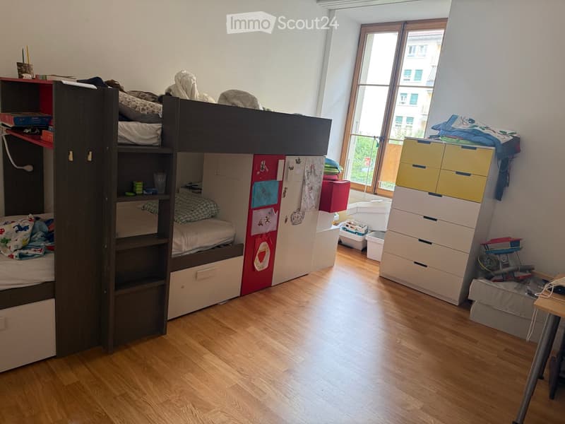 A well-lit bedroom with a lofted twin-sized bed, a wardrobe, a storage unit, and a small table with a chair.
