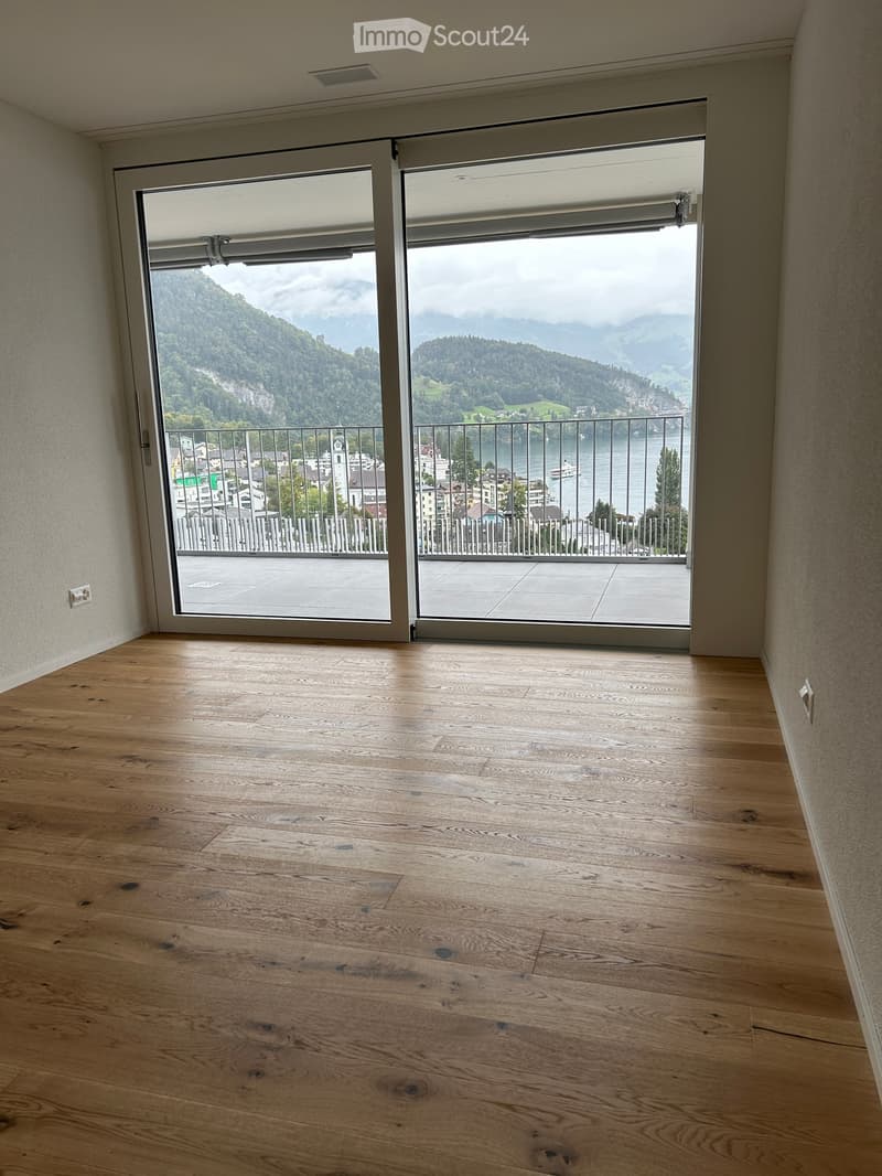 Empty room with wooden flooring and large sliding glass doors opening to a balcony with scenic mountain and lake views.