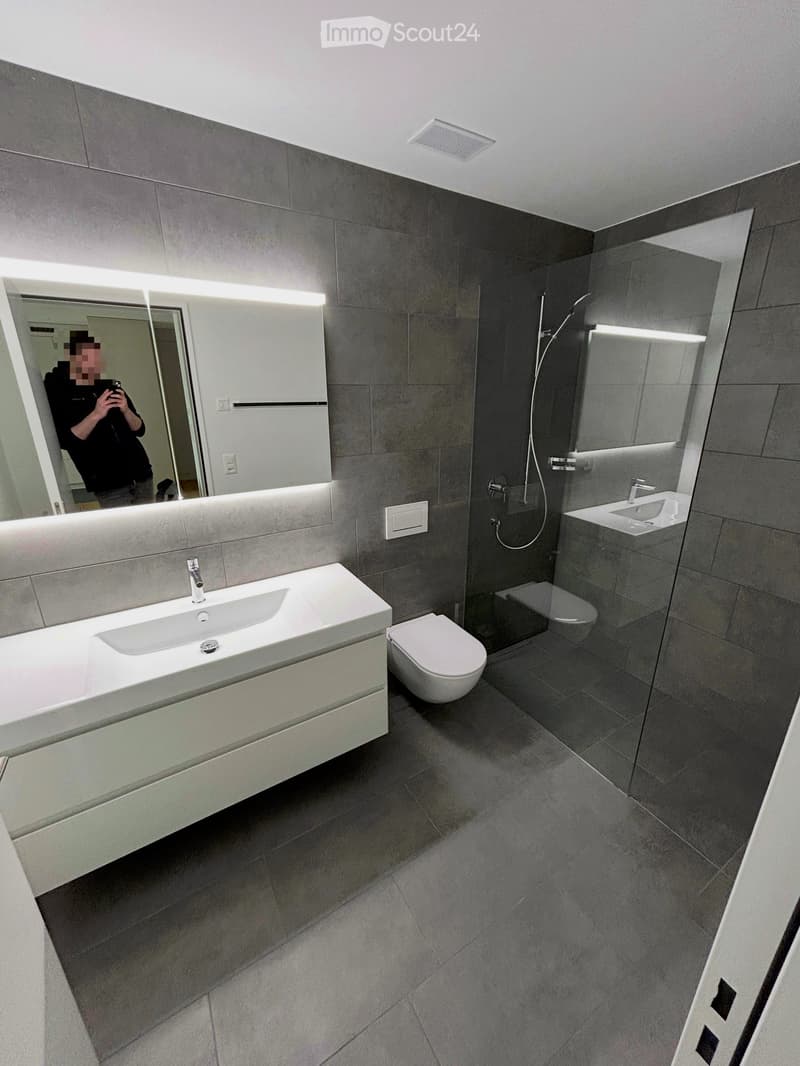 Modern bathroom with grey tiles, white sink, toilet, and large mirror with built-in lighting.
