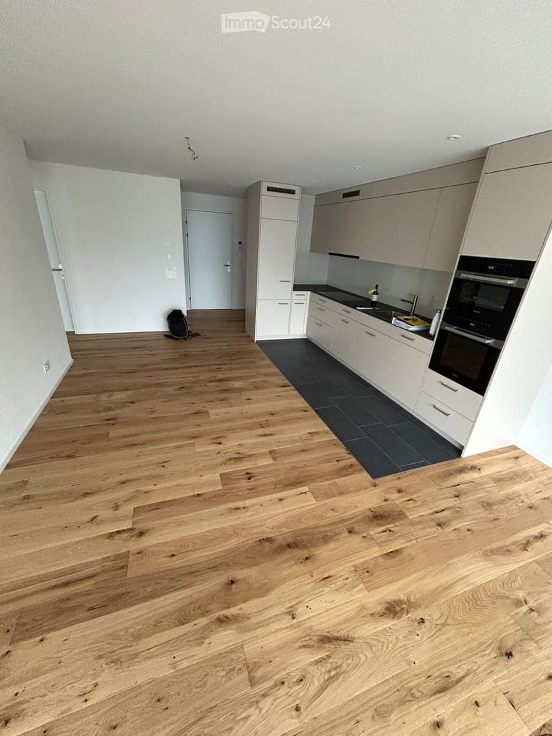 Empty room with light wooden flooring, white walls, and a small kitchen area with white cabinets and black countertops.
