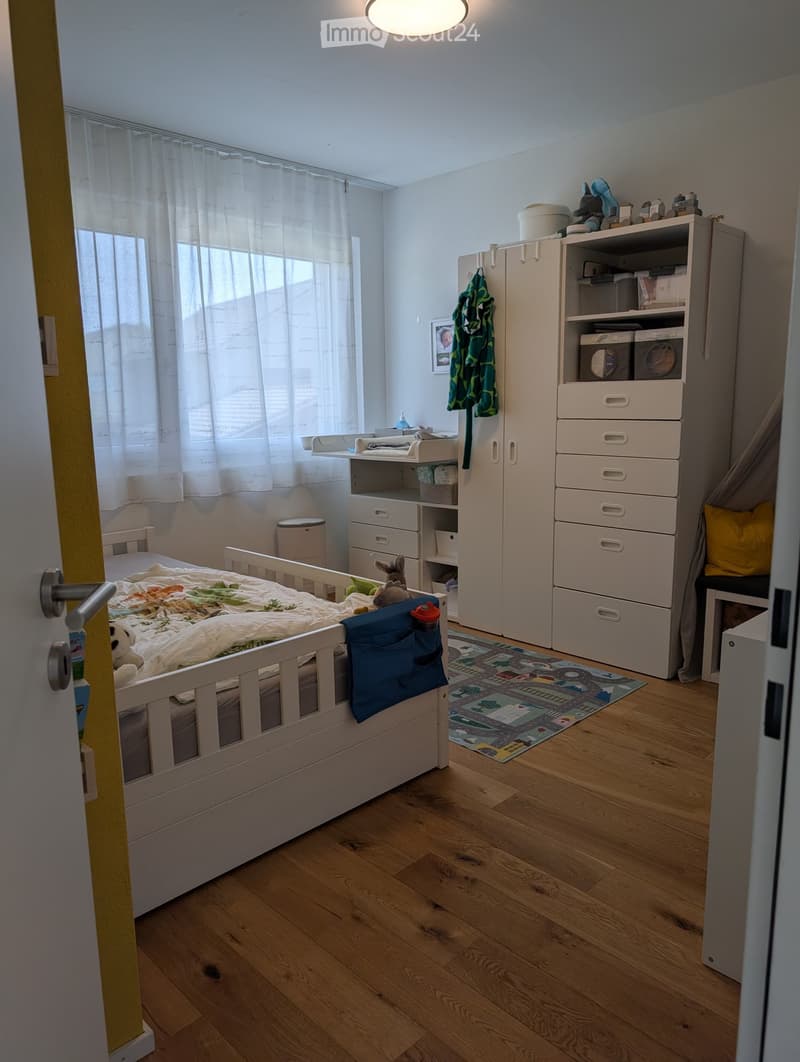 A well-lit children's bedroom with a white wooden bed, a wardrobe, a shelf with storage boxes, a small stool, a colorful play mat, and various toys.