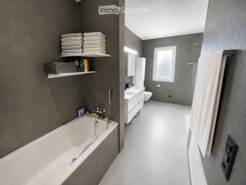 Modern bathroom with dark grey walls, white bathtub, sink, toilet, and shelves holding towels and toiletries.
