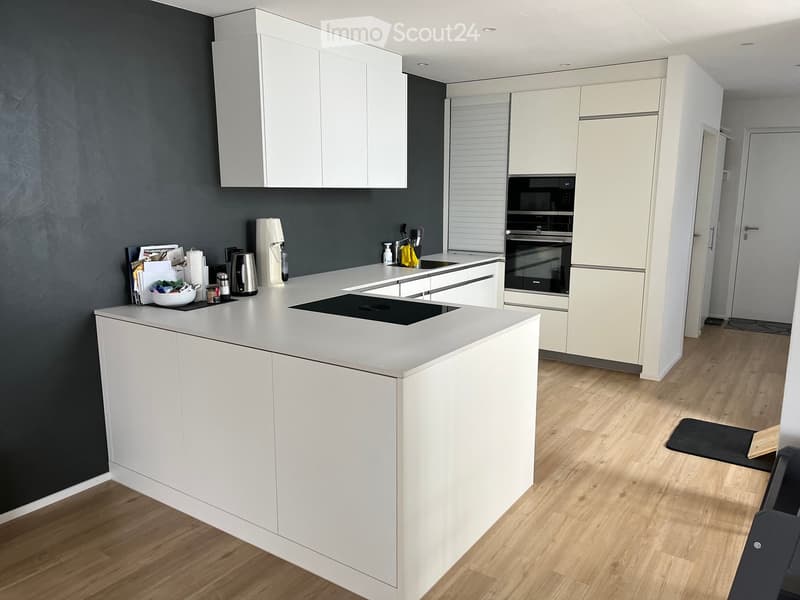 Modern kitchen with white cabinets, a central island, and built-in appliances including an oven and refrigerator. The countertop features a coffee maker, a bowl, and other small items.