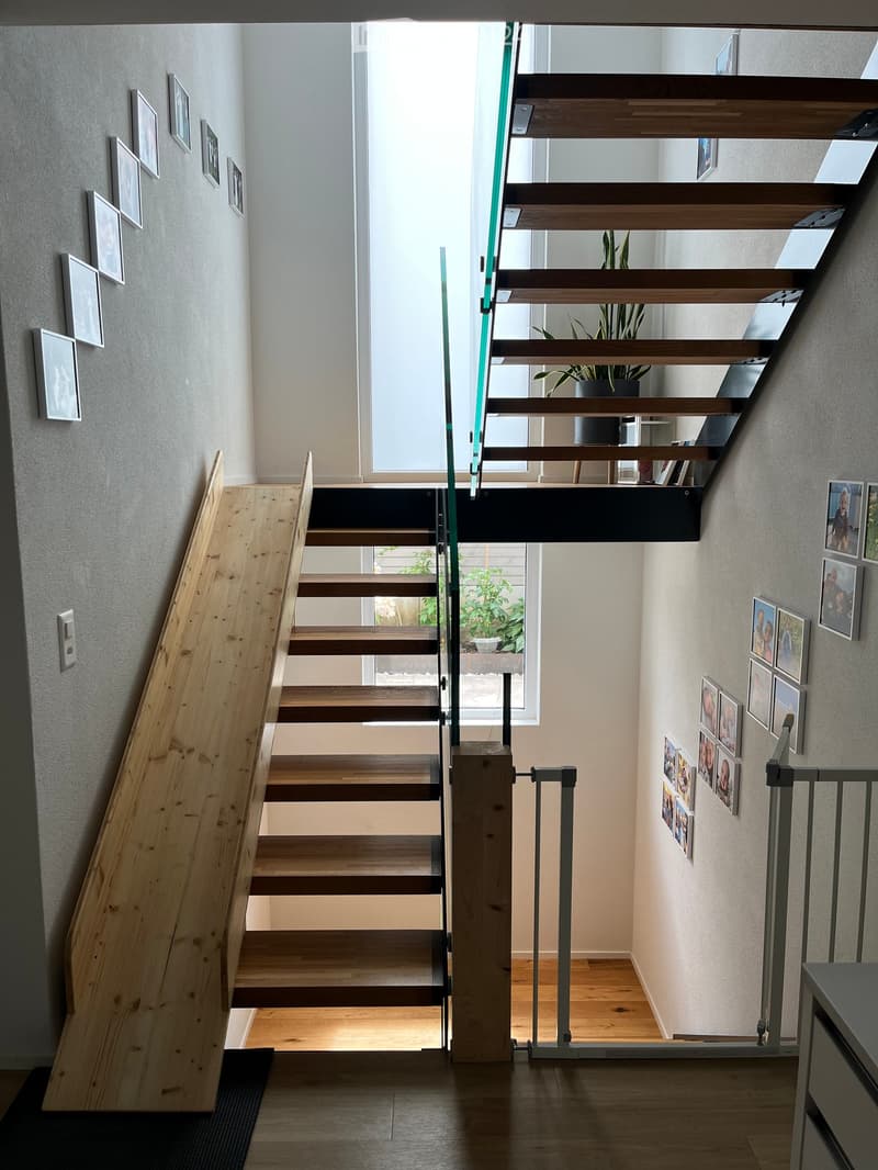 Modern hallway with wooden staircase, glass railing, and framed photos on walls. Features a small wooden slide and a safety gate.