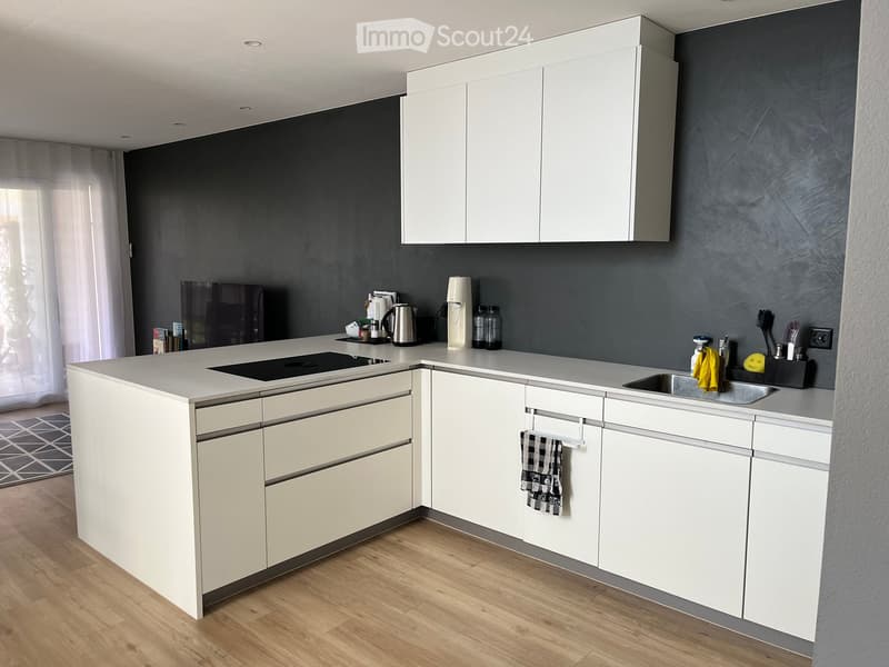 Modern kitchen with white cabinets, grey walls, and a light grey countertop. Features include an electric kettle, coffee maker, sink, and various small appliances.