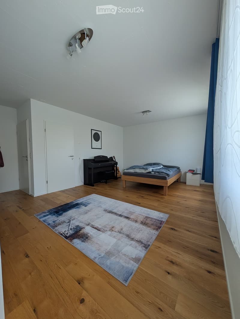 Bright bedroom with wooden flooring, a bed with blue bedding, a piano, a guitar, a nightstand, and a large abstract rug.