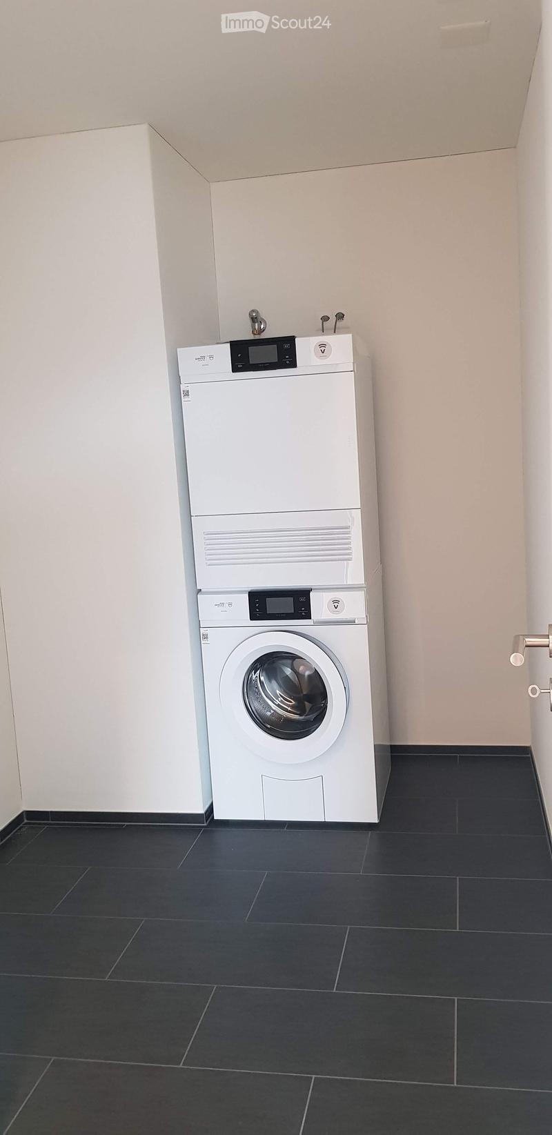 A room with a white washing machine and a white dishwasher stacked on top of each other, against a white wall, with black tiled floor.