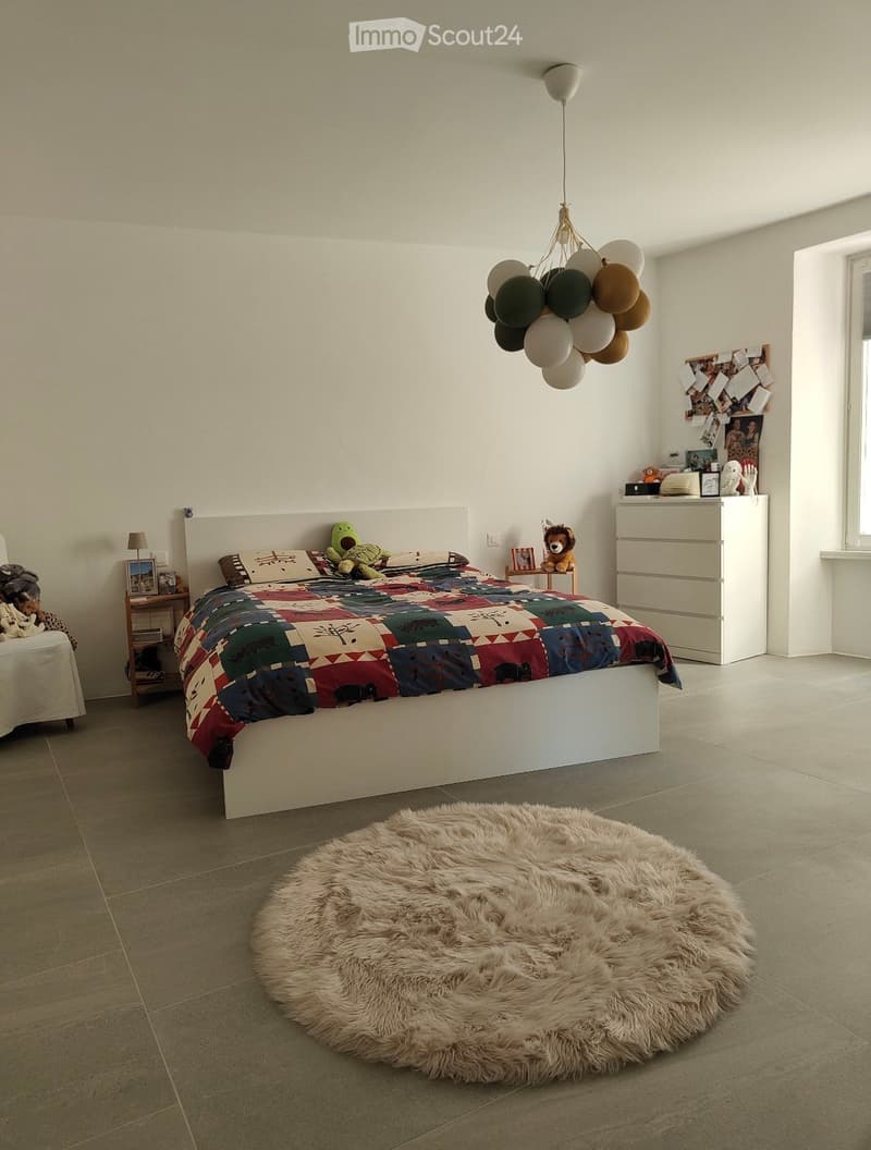 A modern bedroom with a large bed, colorful patchwork bedspread, a round furry rug, a balloon chandelier, and a nightstand with a lamp and toys.