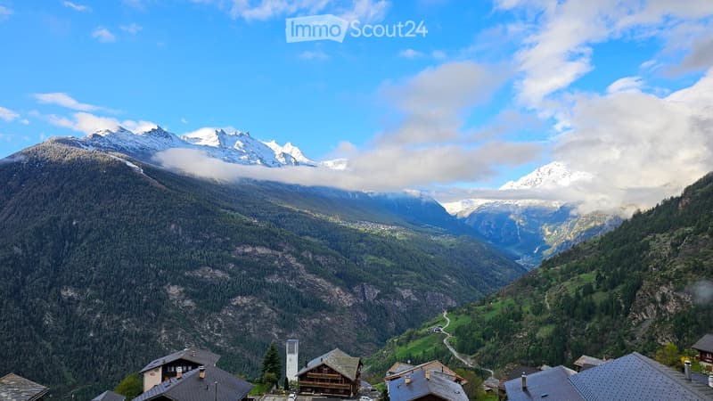 A panoramic view of a mountain valley with snow-capped peaks, dense forests, and scattered houses in the foreground.