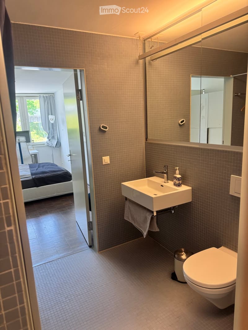Modern bathroom with a white sink, toilet, large mirror, and towel. The adjacent bedroom is visible through an open door, featuring a bed with dark bedding and a window with curtains.