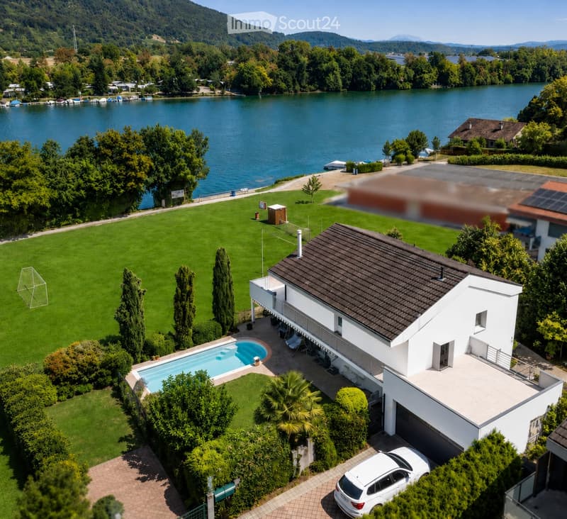 A modern two-story house with a dark roof and white walls, situated near a lake with a swimming pool, lawn, and surrounding greenery.