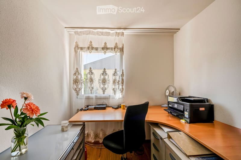 A well-lit home office with a wooden desk, black office chair, printer, fan, and a vase with flowers on a glass table.