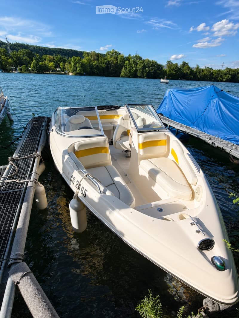 A white boat with beige and yellow seating is docked at a marina, with a calm lake and forested area in the background.