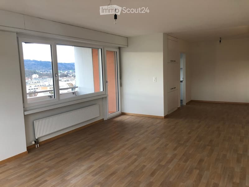 Empty room with wooden flooring, white walls, large window with view, radiator, and a sliding door leading to a balcony.