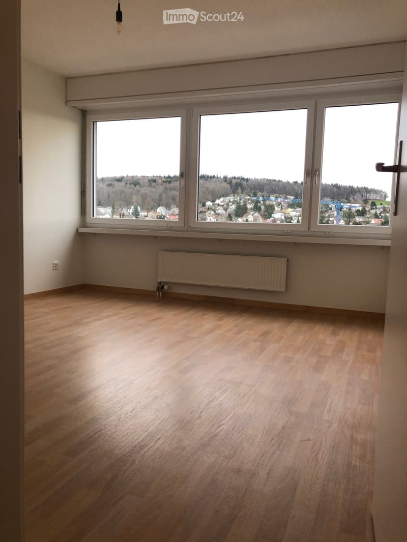 Empty room with wooden flooring, large windows offering a view of a town and surrounding landscape, radiator, and a hanging light fixture.