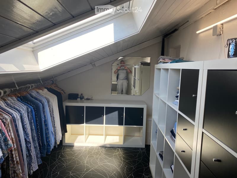 Well-lit walk-in closet with white built-in wardrobe, clothing rack, and large mirror. Features modern design with dark flooring and ample storage space.