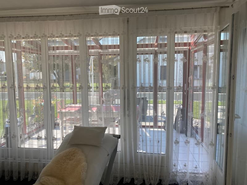 A living room with a sofa and a pillow, large windows with sheer curtains, and a view of an outdoor patio with furniture.