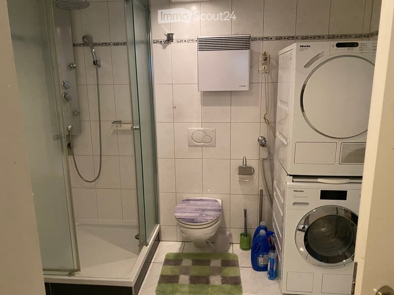 Modern bathroom with a glass-enclosed shower, toilet, washing machine, and cleaning supplies on the floor.