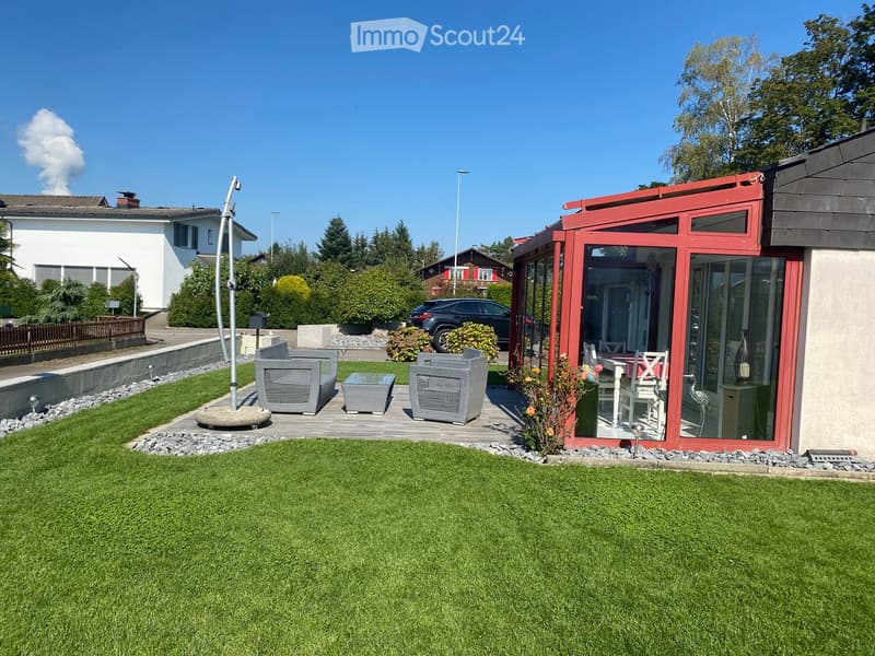 Well-maintained garden with a wooden deck, grey outdoor sofa, coffee table, dining table, chairs, and decorative vase inside a red summerhouse.