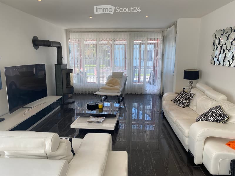 Modern living room with white leather sofa, coffee table, fireplace, large windows with sheer curtains, and a television on a stand.