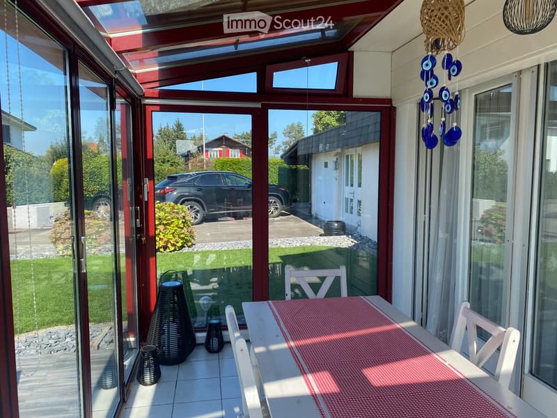 Bright, enclosed patio with white table, red tablecloth, white chairs, and decorative wicker lamps. Large glass doors open to a well-maintained garden and driveway with a parked car.