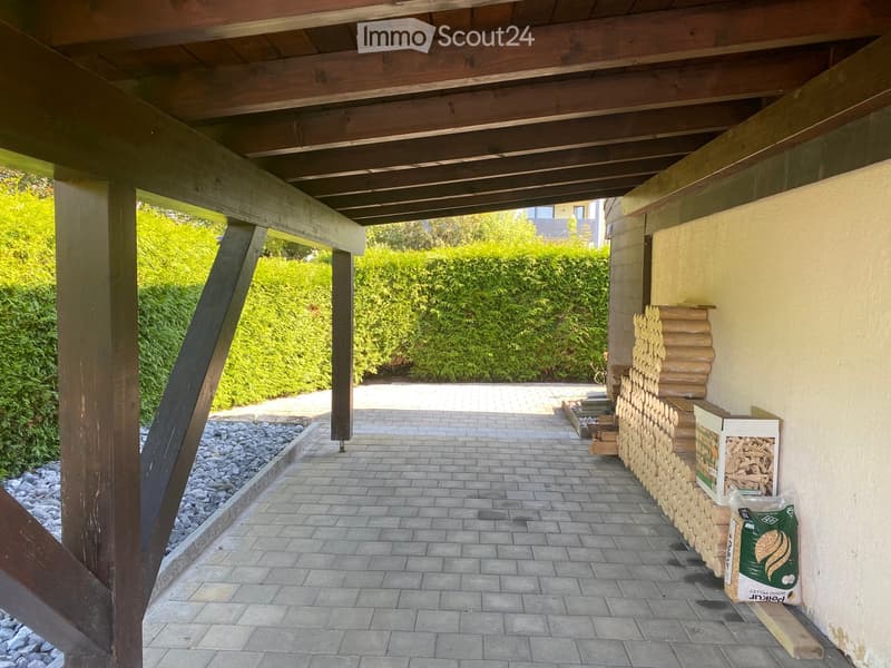 Wooden covered outdoor area with brick paved floor, stacked firewood, and surrounding greenery.