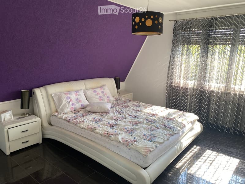 A modern bedroom with a white upholstered bed, floral bedding, nightstands with lamps, and large windows with sheer curtains.