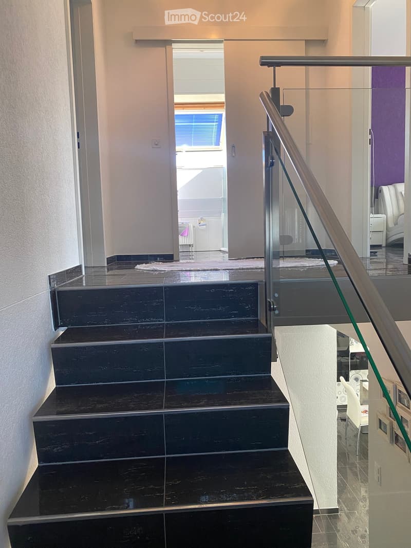 Modern staircase with dark wooden steps and a glass railing, leading to a bright room with white furniture and a purple accent wall.