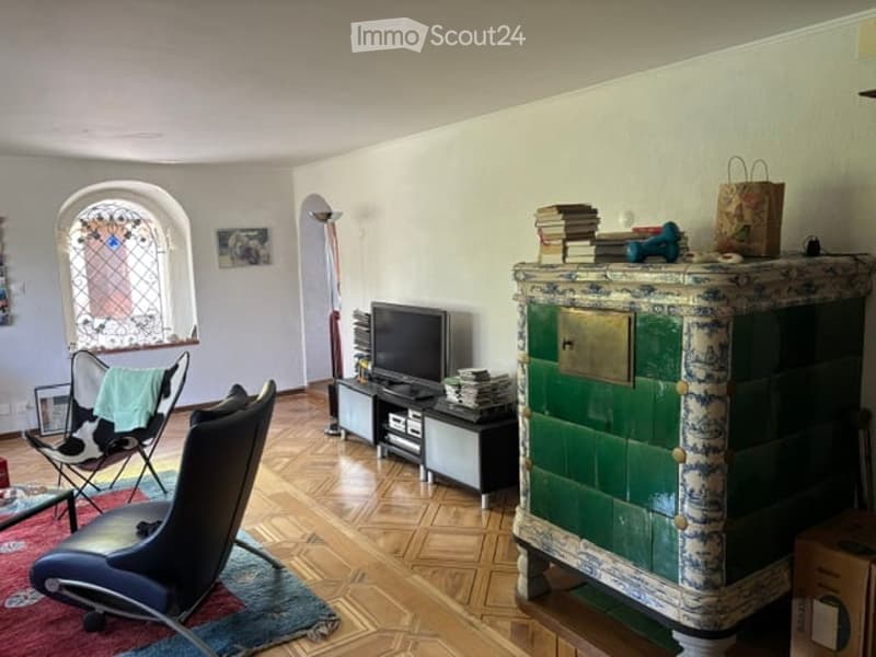 Living room with wooden flooring, large window with stained glass, green and white painted cabinet, sofa, chair, television, and books on top of the cabinet.