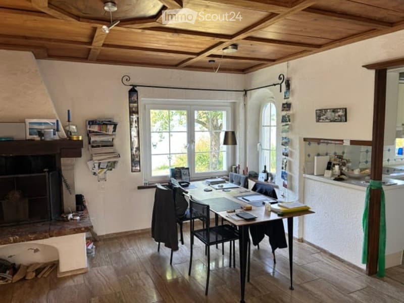 Bright room with wooden ceiling beams, fireplace, large windows, and a dining table with chairs.