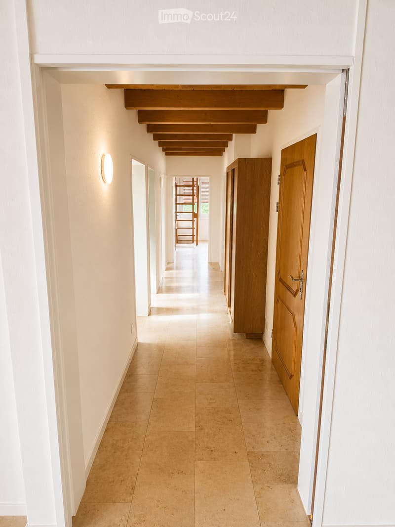 Clean, modern hallway with beige tiled flooring, white walls, and wooden doors. A staircase with wooden railings is visible in the background, along with a wall-mounted light fixture on the left side.