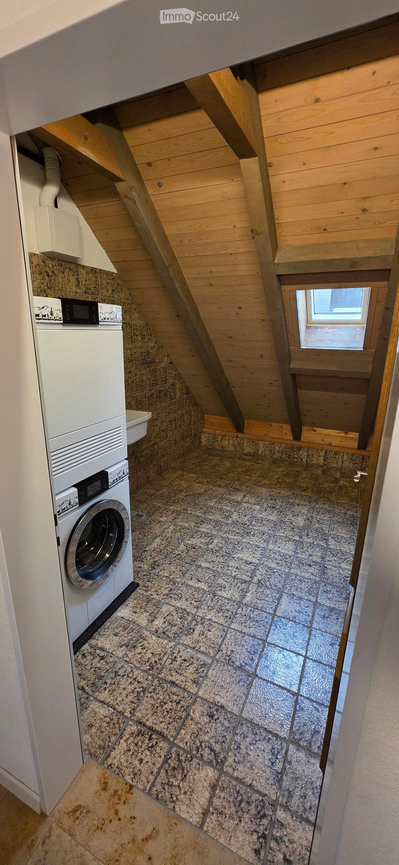 Attic bathroom with a washing machine, granite tiled floor, wooden ceiling beams, and a small window.