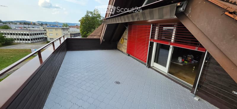 Balcony with wooden railing, grey tiled floor, red shutters, and an open door leading to a small storage area.