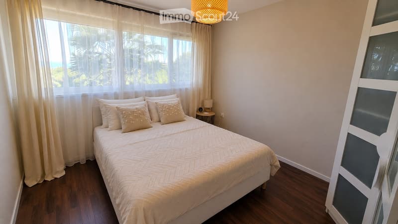 A modern bedroom with a large bed, multiple pillows, a nightstand with a lamp, and large windows with sheer curtains offering a view of palm trees and the ocean.