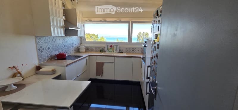 Bright kitchen with white cabinets, black tiled floor, and a large window offering a sea view. Includes a refrigerator, sink, oven, countertop, and various kitchen items.