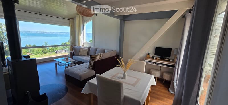 Bright living room with large windows offering a scenic view, featuring a sofa, coffee table, ottoman, dining table, chairs, television, and a wicker lamp.