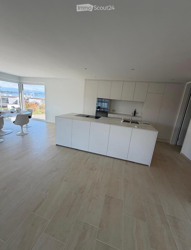 Modern kitchen with white cabinets, a central island, and a large window overlooking the ocean. The floor is tiled with a mix of light and dark tiles.