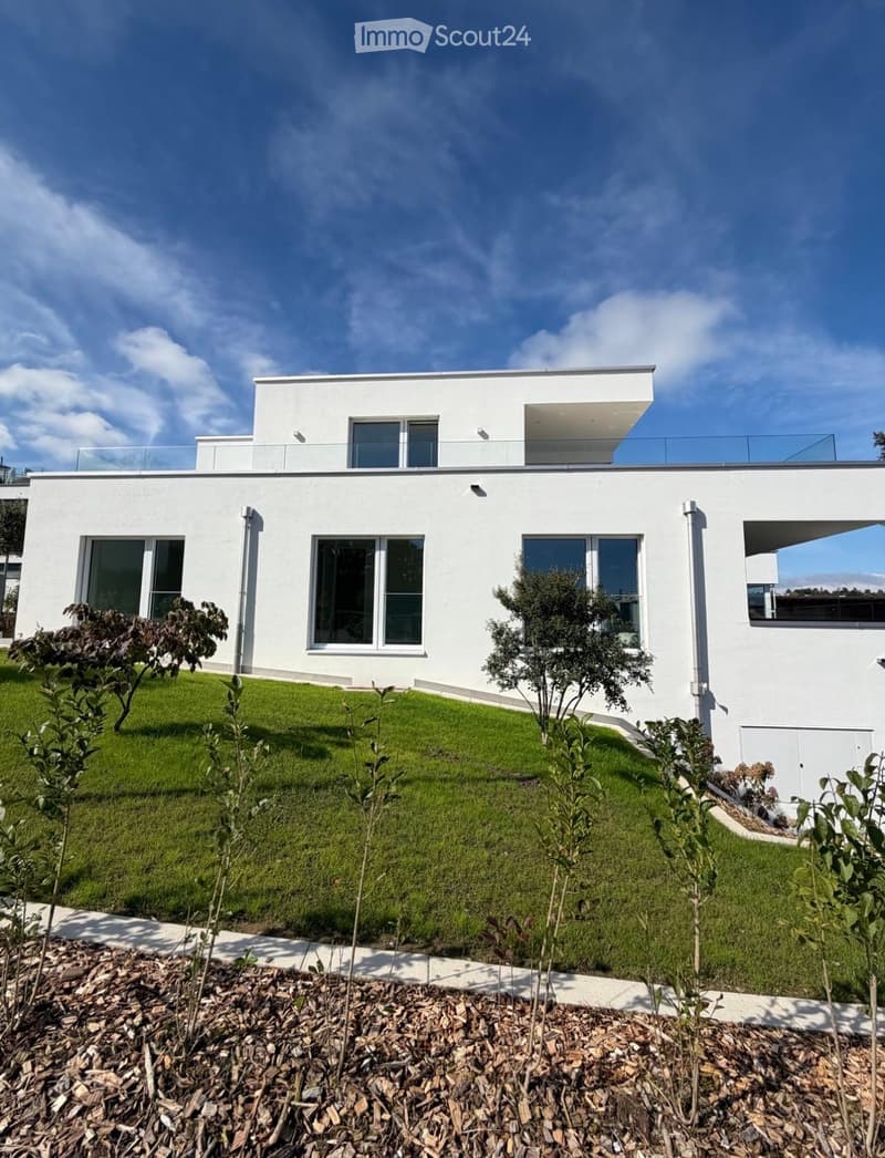 Modern two-story white house with large windows, a flat roof, and a raised ground floor. The house features a well-maintained lawn and small trees in the front yard.