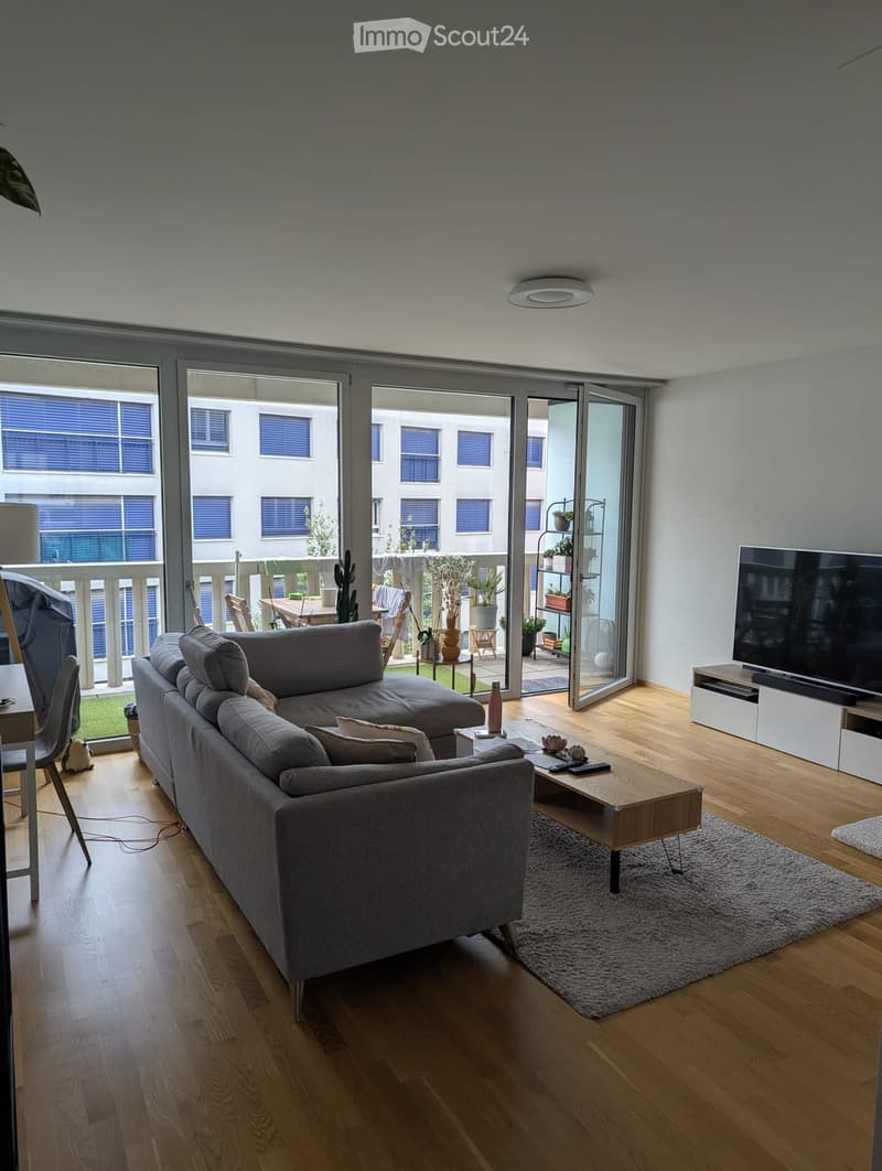 Modern living room with wooden flooring, grey sectional sofa, coffee table, TV, and access to a balcony with outdoor furniture.
