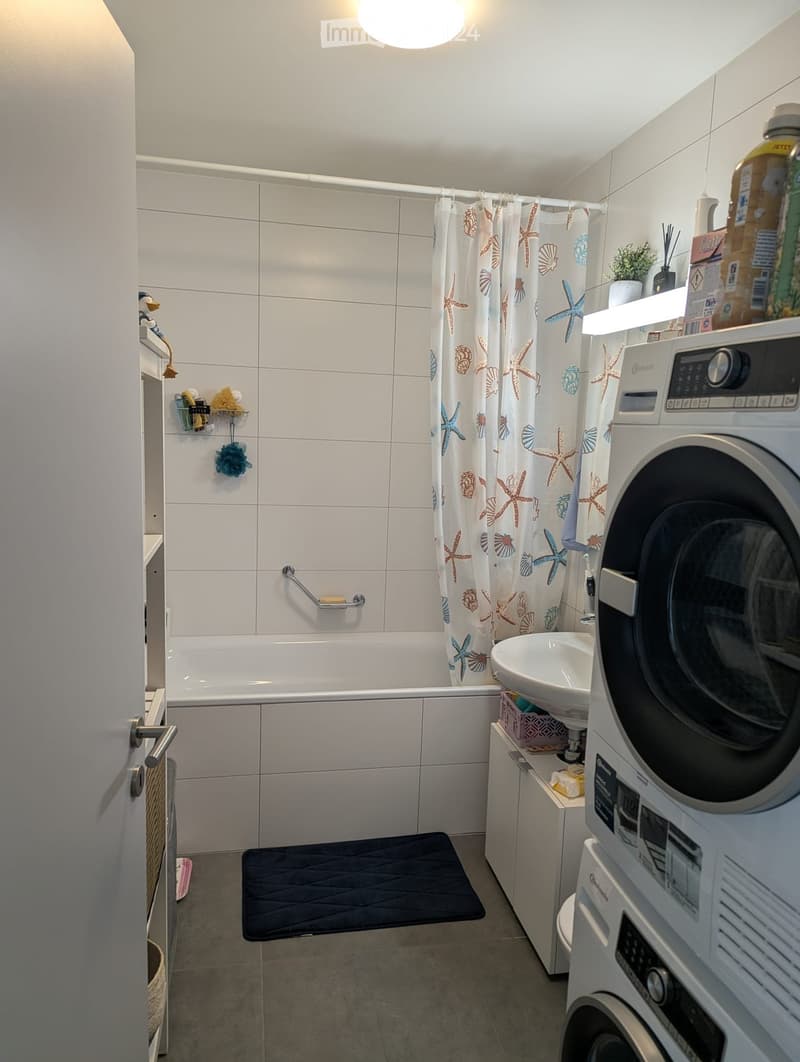 Modern bathroom with a bathtub, shower curtain, sink, washing machine, and a small shelf with toiletries.