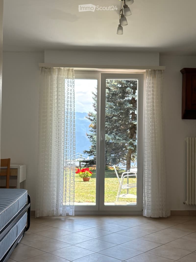 Bright bedroom with tiled floor, large window with view of mountains, bed, chair, and sheer curtains.