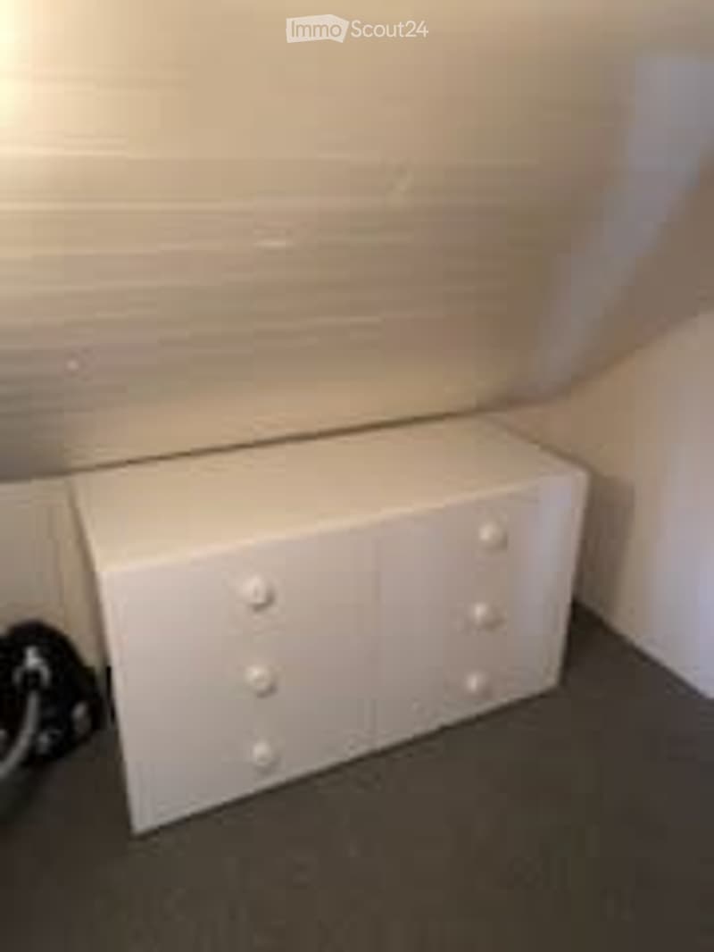 An empty room with white walls and a white cabinet with four drawers and four knobs. The floor is carpeted in dark grey.