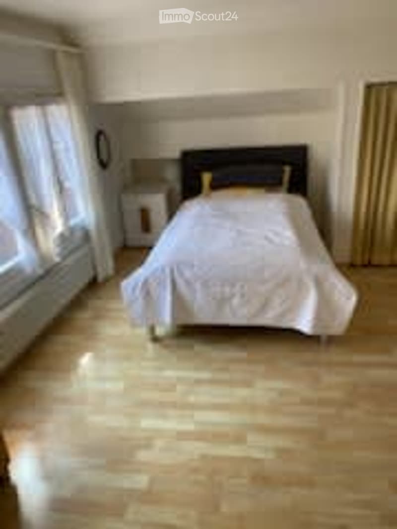 Bright bedroom with wooden flooring, large windows, a neatly made bed with white bedding, a nightstand, and a lamp.